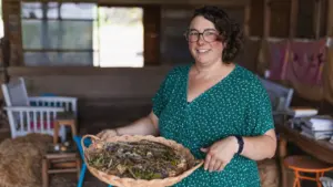 Une femme souriante aux cheveux courts porte une robe verte tenant un plateau d'herbes séchées.