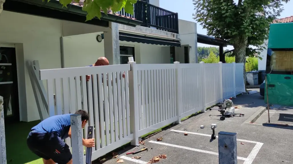Deux ouvriers installent une clôture en PVC blanc devant une maison résidentielle par beau temps.