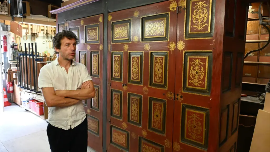 Un ébéniste debout devant une imposante armoire ancienne en bois rouge avec des détails dorés.