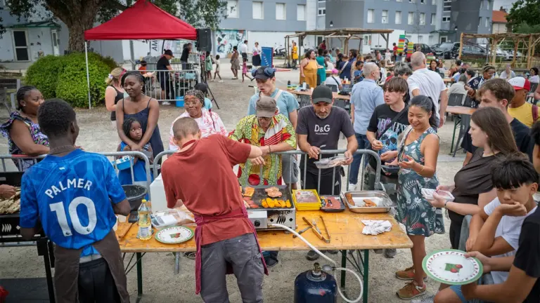Un homme cuisine des grillades sur une table en extérieur devant une foule attentive.